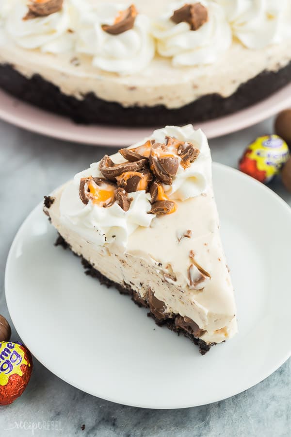 slice of no bake creme egg cheesecake on white plate with whipped cream and crushed creme eggs
