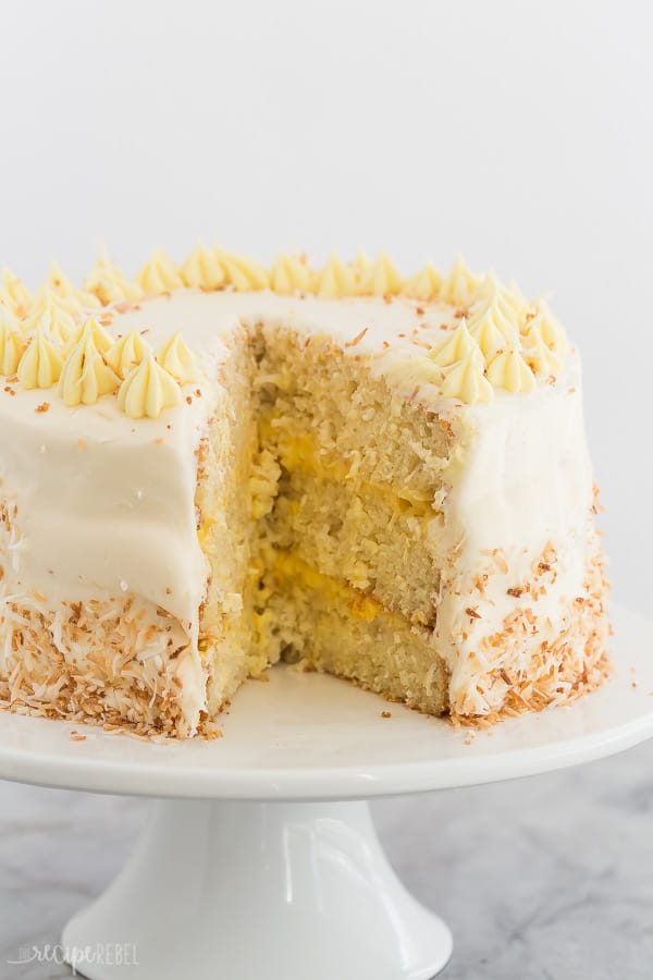 This Pineapple Coconut Cake is light, fresh and fruity, and covered in cream cheese whipped cream! It's the perfect Easter dessert or anytime treat! With step by step VIDEO. #recipe #dessert #cake #pineapple #coconut #easter pineapple coconut cake whole with cream cheese frosting and one slice cut out