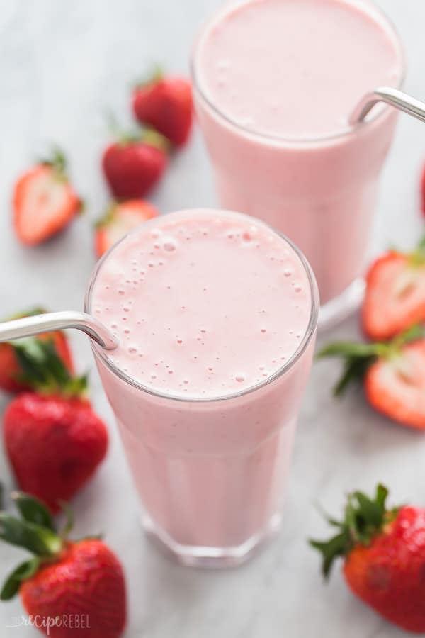 Two tall glasses of pale pink strawberry smoothie, with stainless steel straws. Fresh strawberry halves decorate the white table. 