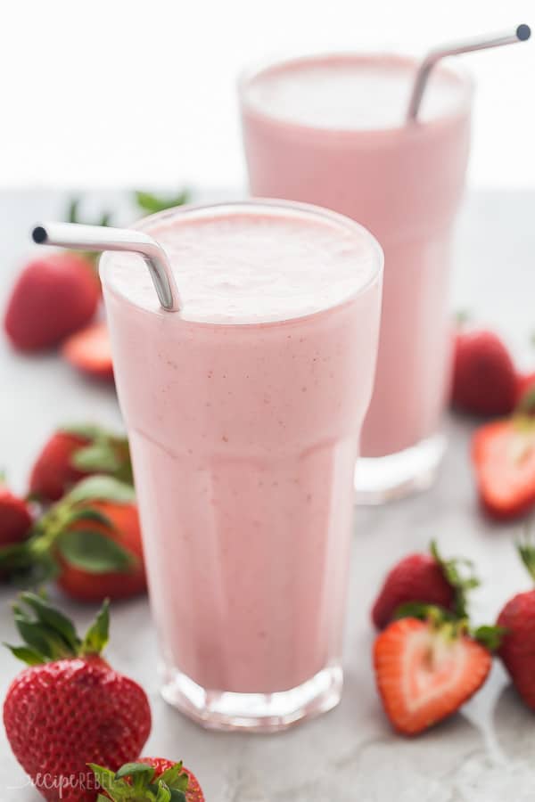 Two strawberry smoothies in glasses with steel straws, on a grey marble background scattered with fresh berries.