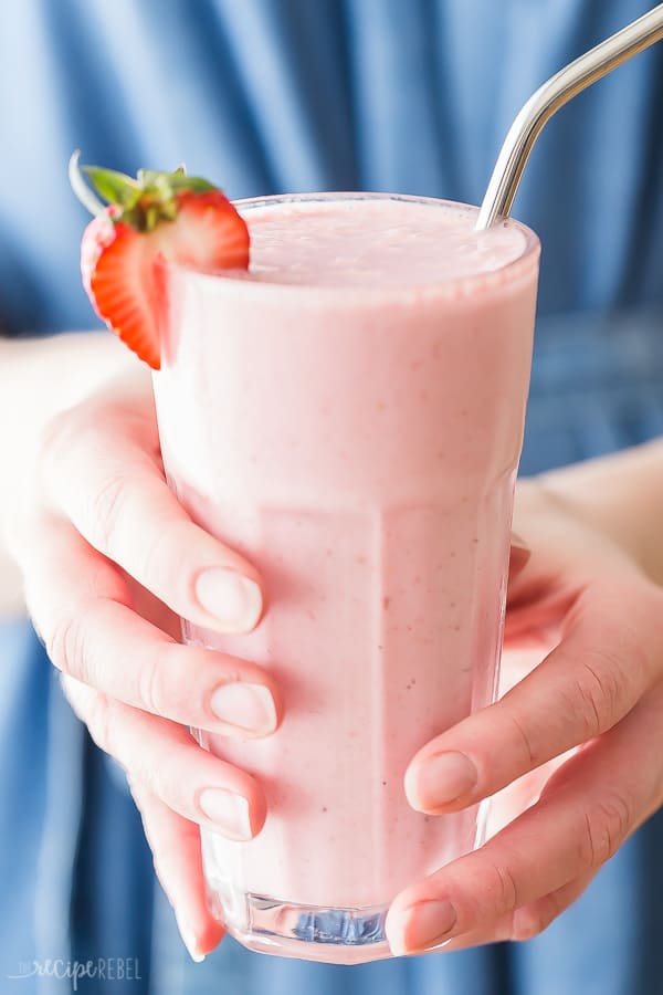 Hands holding a glass filled to the top with pink smoothie. The glass is garnished with half a strawberry.