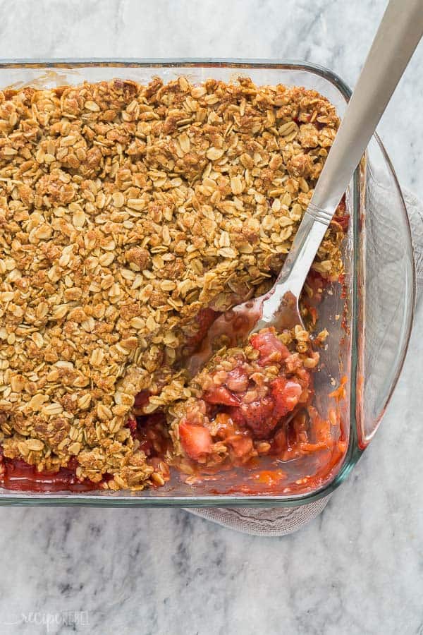 strawberry rhubarb crisp in pan on marble background with metal spoon scooping