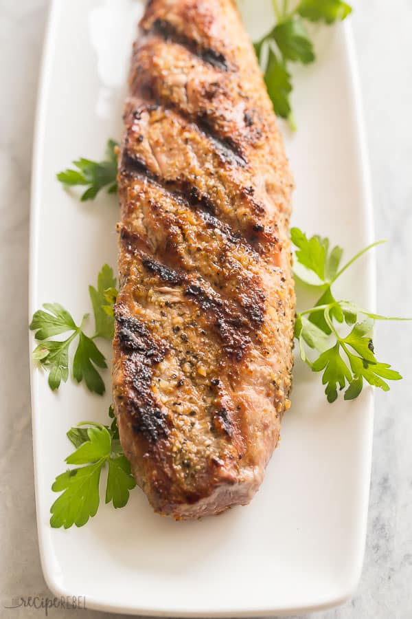 grilled pork tenderloin whole on white plate with sprigs of fresh parsley