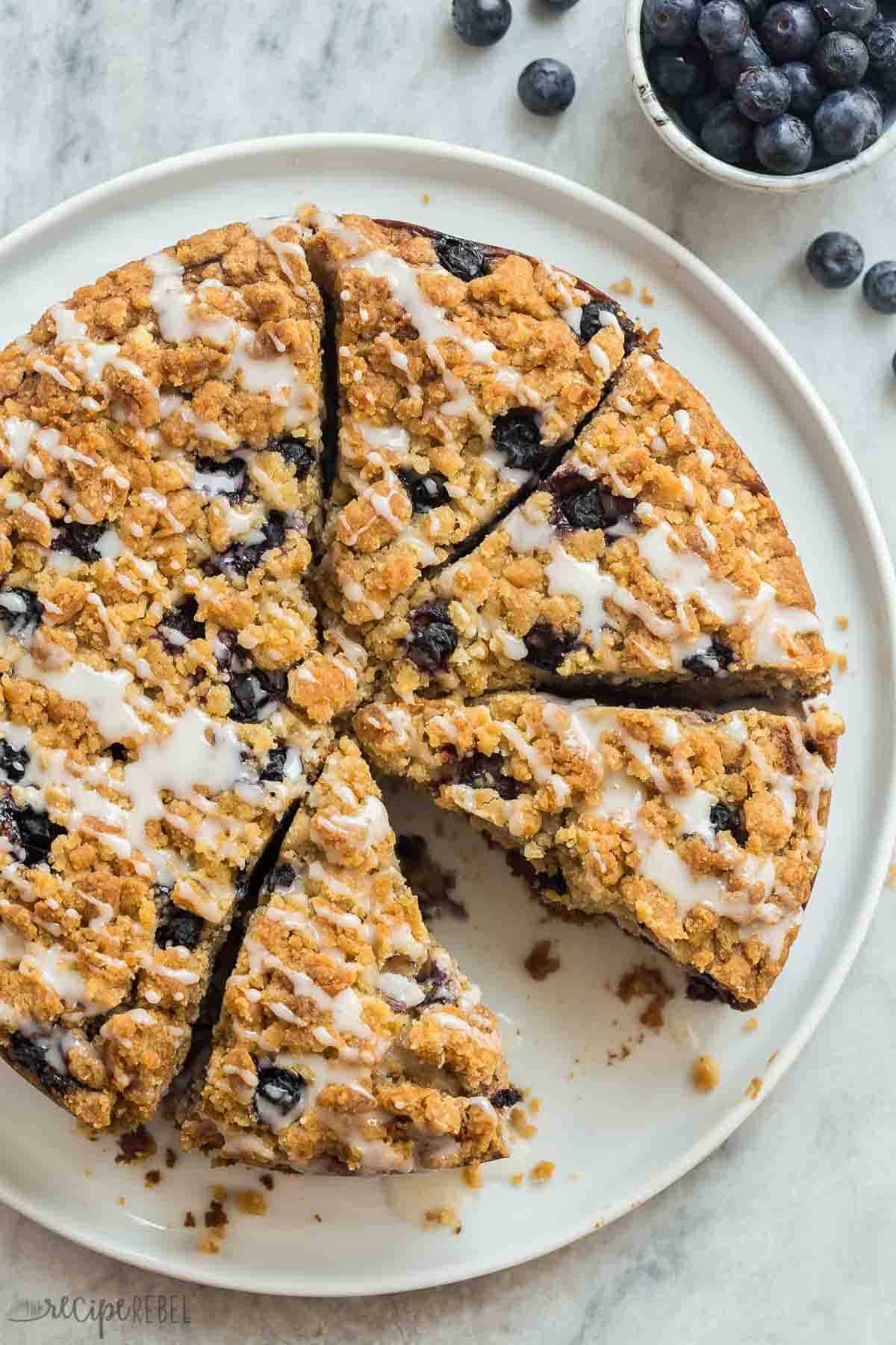 This Blueberry Coffee Cake is moist and loaded with juicy blueberries and crunchy brown sugar streusel -- it's perfect for brunch or dessert and uses fresh or frozen berries! #blueberry #blueberries #cake #recipe #brunch | coffee cake recipe | easy baking recipes | brunch goals | blueberry recipes | easter brunch overhead image of a blueberry coffee cake cut into slices on a white plate.