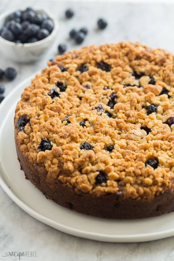 blueberry coffee cake whole on white plate with fresh blueberries in the background