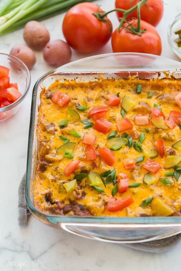 This easy Cheeseburger Casserole is an easy dinner idea that is make ahead and freezer friendly, loaded with ground beef, potatoes, cheese, and all your favorite cheeseburger flavors! #groundbeef #beef #potatoes #recipe #dinner cheeseburger casserole in glass pan with pickles tomatoes and green onions on top