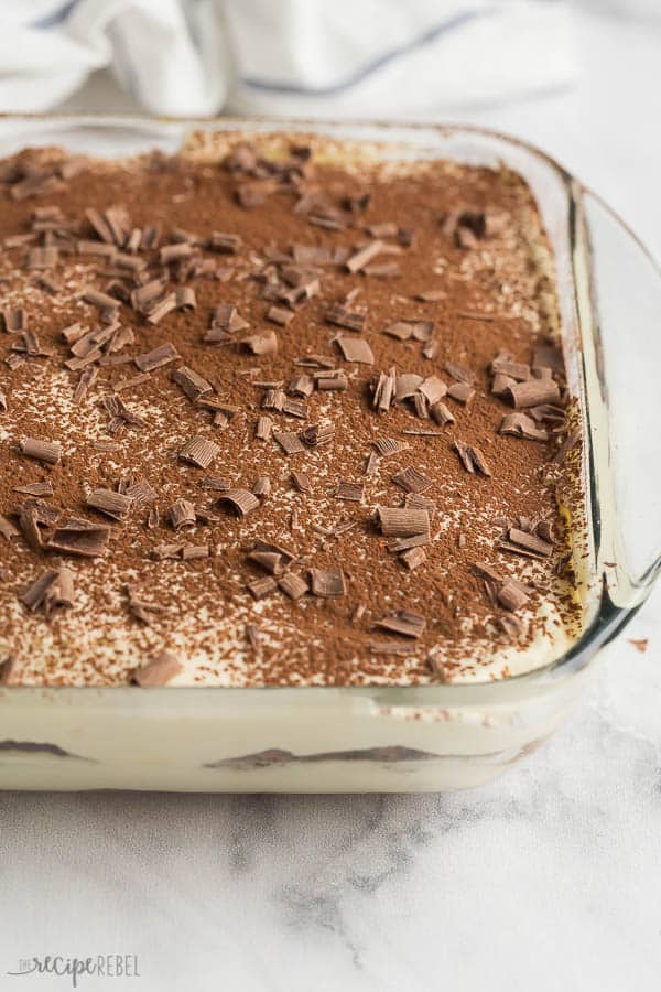 This easy Tiramisu recipe is made with a traditional mascarpone custard filling, coffee soaked ladyfingers, and a fine dusting of cocoa powder and chocolate shavings. It's an easy no bake dessert! #recipe #dessert #coffee #dessertrecipe #nobake tiramisu pan whole in glass pan with chocolate shavings on marble background