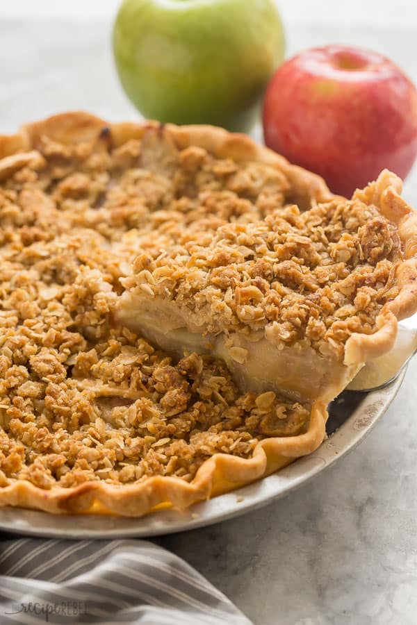 apple crumble pie slice being pulled out of whole pie with apples in the background