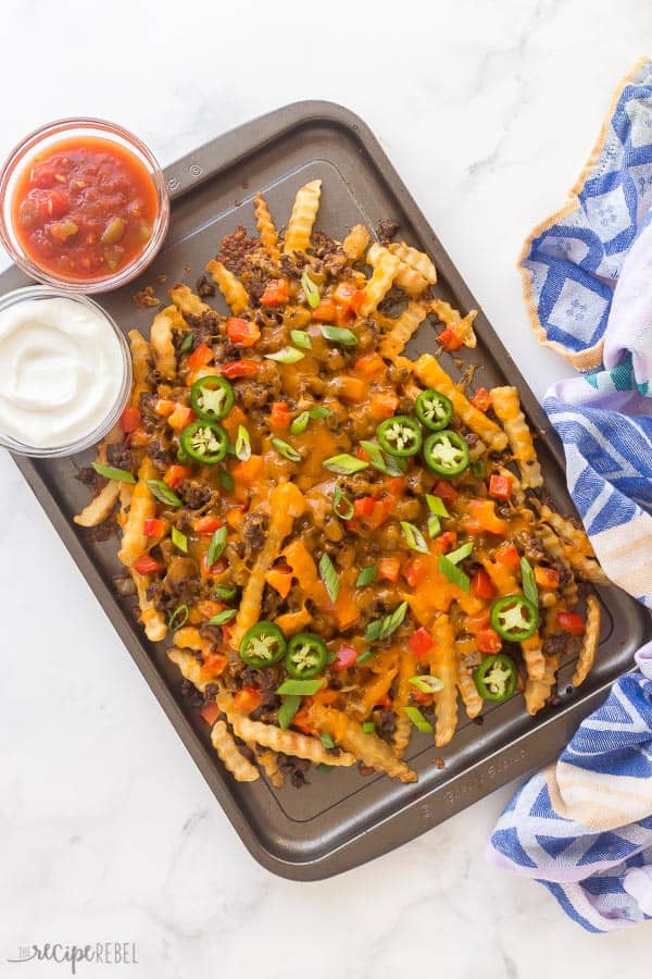 These Nacho Fries are better than Taco Bell! Perfectly seasoned and topped with taco beef, lots of cheese, peppers, and green onions. #fries #appetizer #beef #cheese #recipes nacho fries overhead on baking pan with jalapeno slices, sour cream and salsa on the side