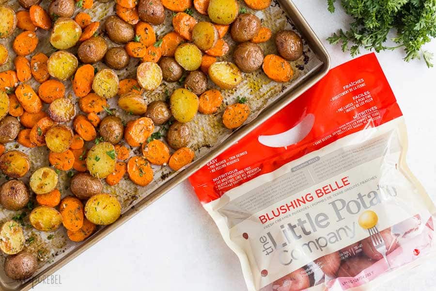 A sheet pan of roasted vegetables on a white work surface. A bunch of fresh parsley is in the upper right corner of the frame, while a bag of The Little Potato Company Blushing Belle Creamer potatoes is in the lower right corner.