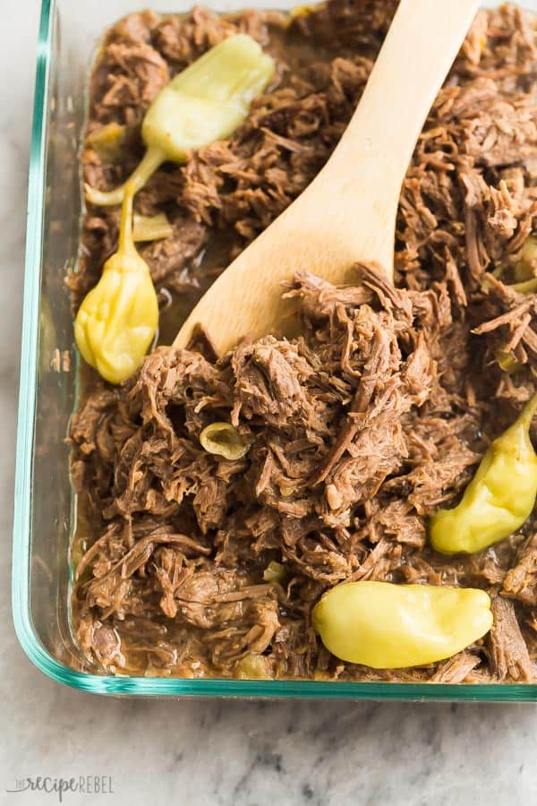 crockpot italian beef in glass dish with pepperoncini on top and wooden spoon