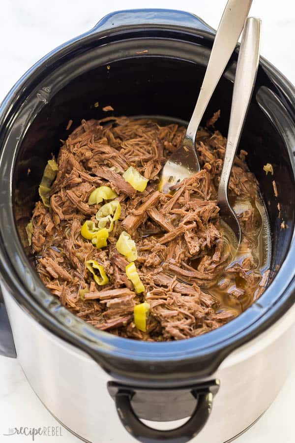 crockpot italian beef in black slow cooker with two forks and peppers on top