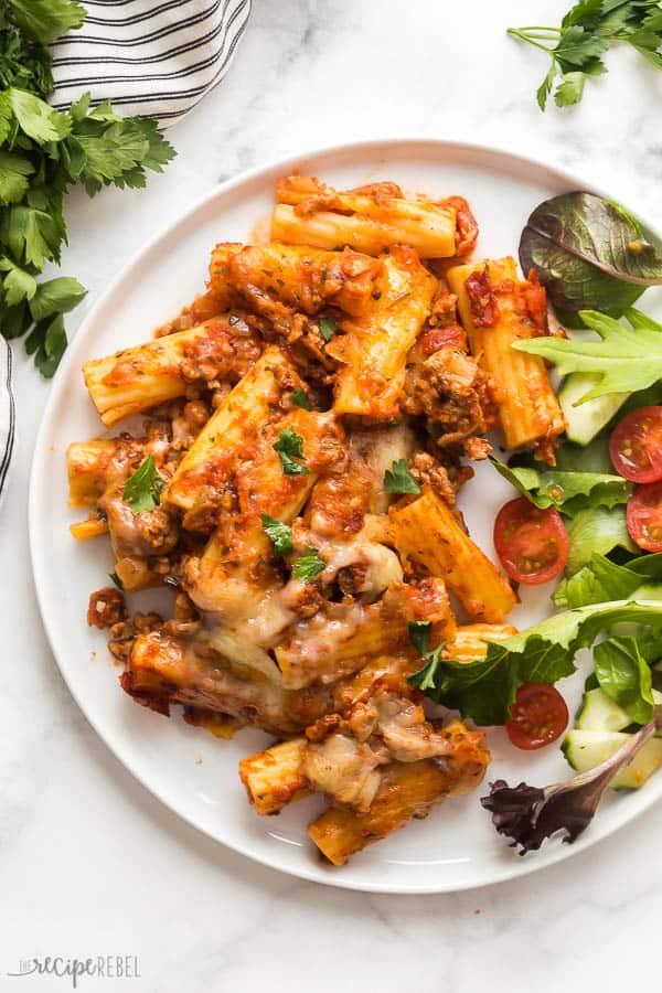 baked ziti on white plate on white marble background with salad