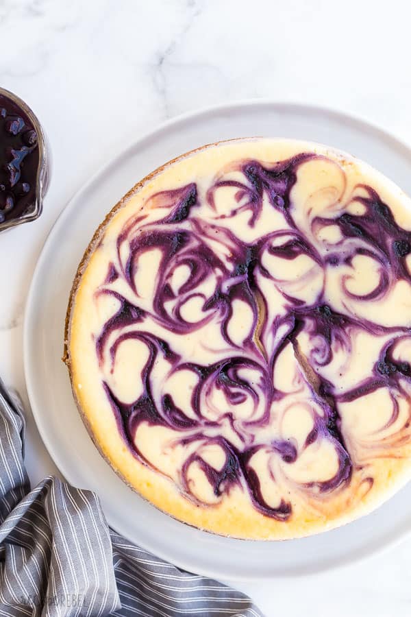 This Blueberry Cheesecake is ultra creamy and swirled with fresh blueberry sauce. It's baked to perfection with no water bath and no fuss! #cheesecake #blueberry #blueberries #dessert #recipe blueberry cheesecake whole on large white plate on marble background