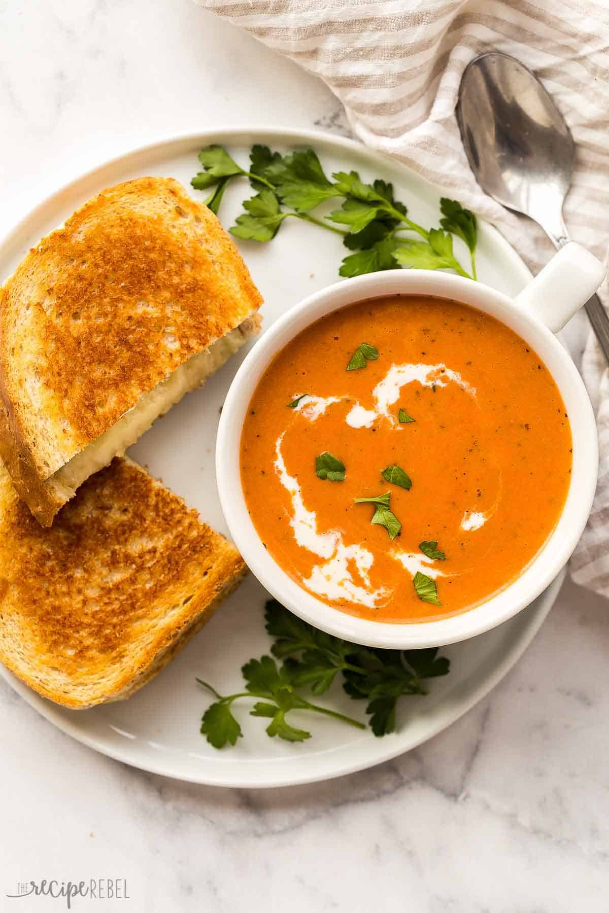overhead image of mug of tomato soup with two pieces of grilled cheese on a plate.