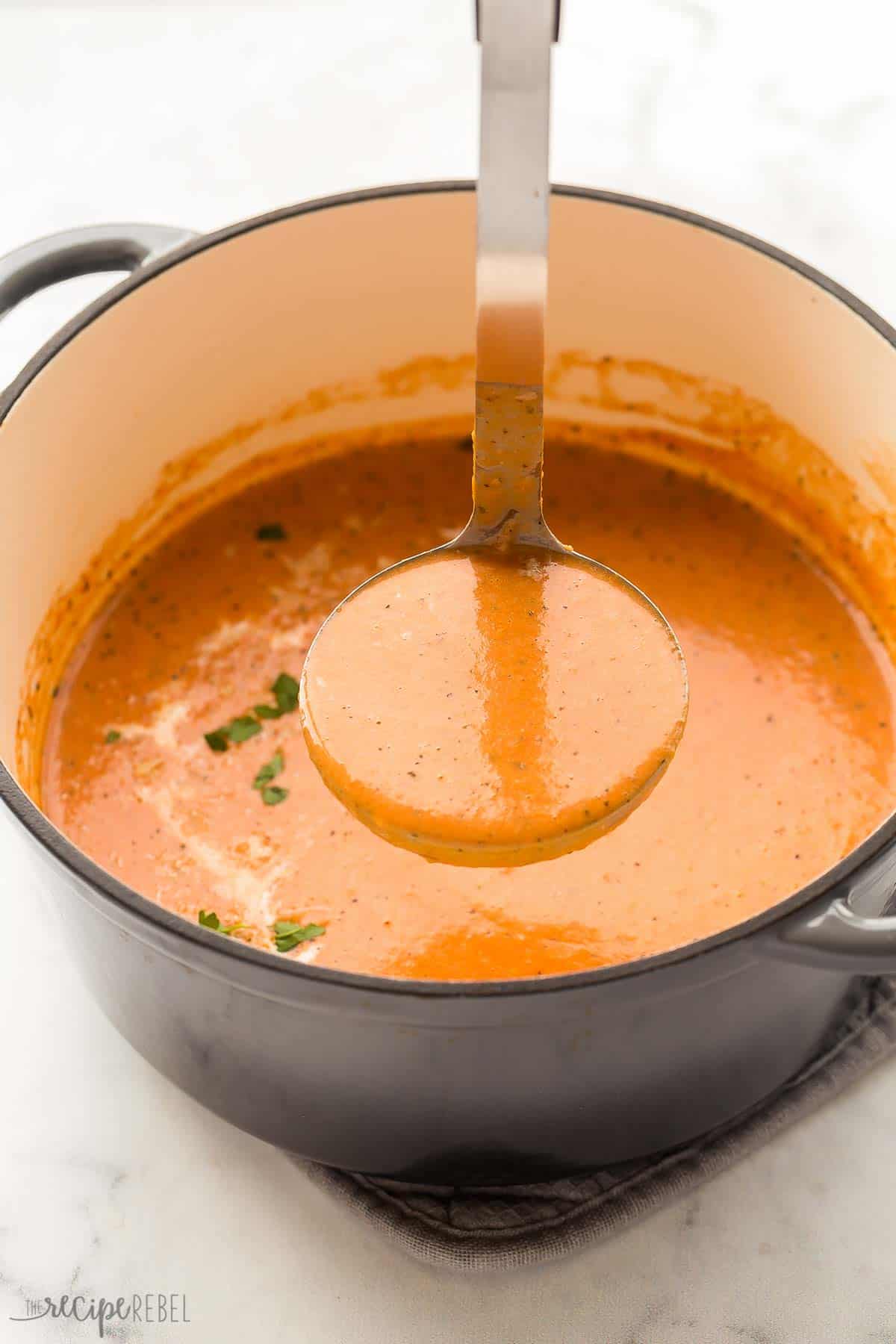 ladle scooping creamy tomato soup out of a grey pot.