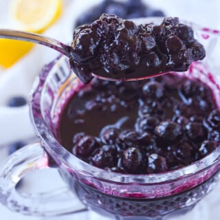 A spoon is used to scoop blueberry compote from a glass serving dish.