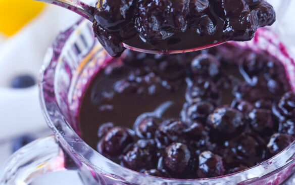 A spoon is used to scoop blueberry compote from a glass serving dish.