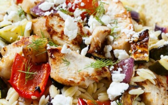 A blue bowl of orzo salad with chicken and roasted vegetables, served with a slice of bread.