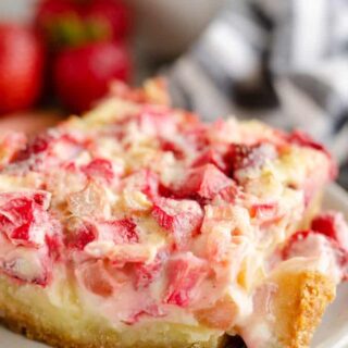 A slice of strawberry rhubarb custard dessert on a plate, with scattered strawberries in the background.