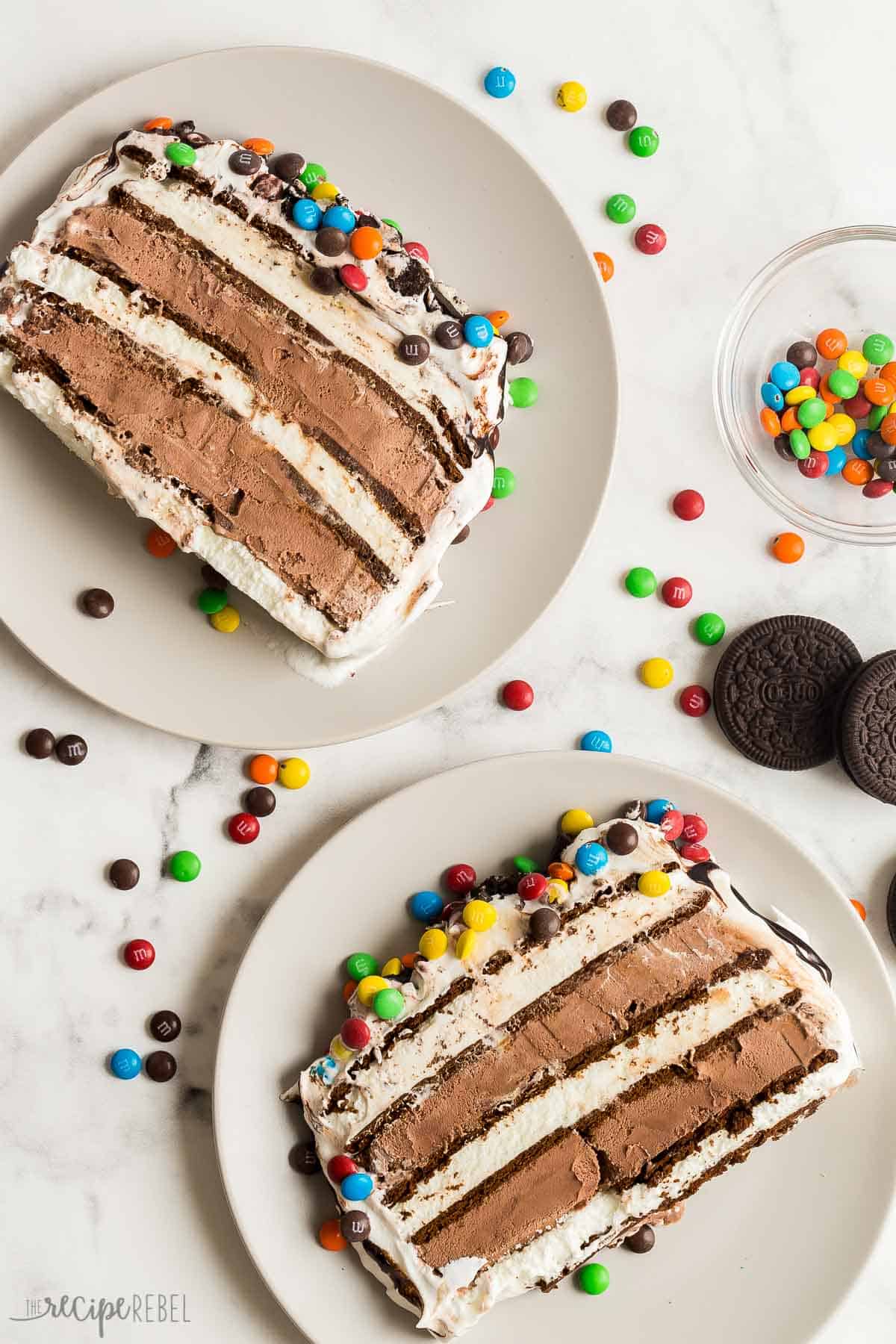 This Ice Cream Sandwich Cake is made with just SIX ingredients or less, no baking required and a ton of easy ways to customize it! Use your favorite store bought ice cream sandwiches and ice cream flavors to make it your own. #dessert #icecream #recipe | ice cream cake recipe | ice cream recipes | easy ice cream dessert | ice cream cake | chocolate ice cream cake | M&Ms | summer dessert | no bake dessert | birthday party | birthday cake | cake recipes | easy dessert overhead image of two slices of ice cream sandwich cake on plates with M&Ms around.