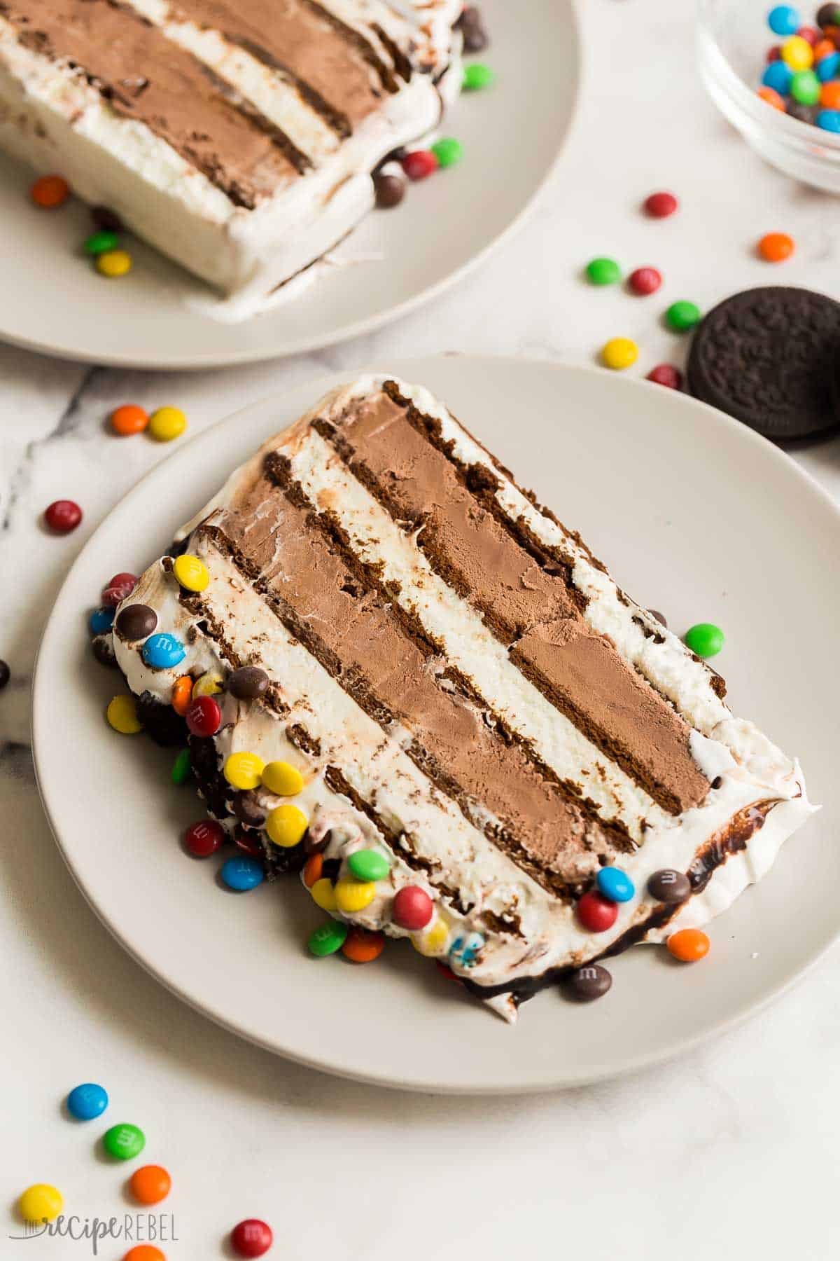 This Ice Cream Sandwich Cake is made with just SIX ingredients or less, no baking required and a ton of easy ways to customize it! Use your favorite store bought ice cream sandwiches and ice cream flavors to make it your own. #dessert #icecream #recipe | ice cream cake recipe | ice cream recipes | easy ice cream dessert | ice cream cake | chocolate ice cream cake | M&Ms | summer dessert | no bake dessert | birthday party | birthday cake | cake recipes | easy dessert slice of ice cream sandwich cake on a plate with mini M&Ms around.