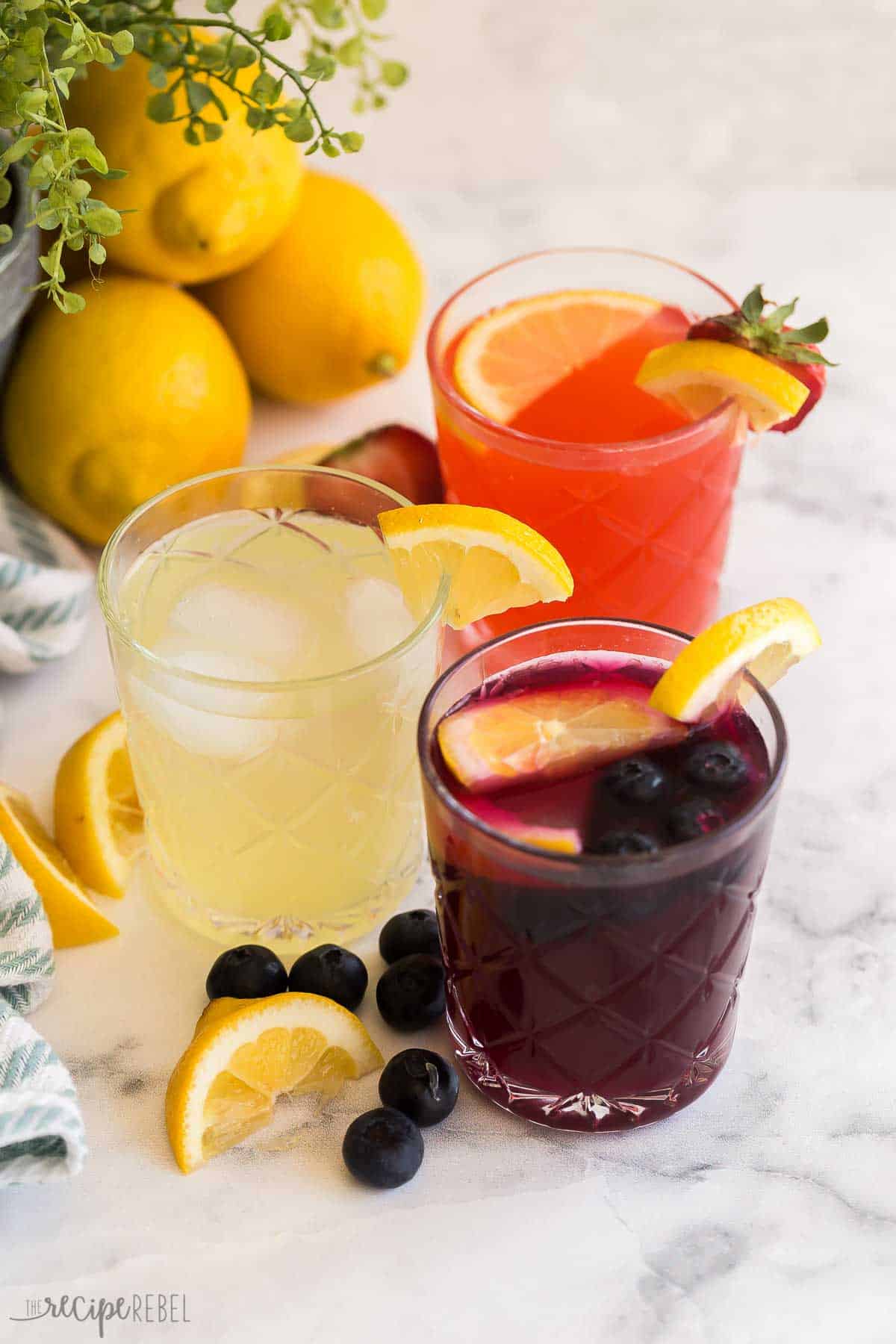 This easy homemade Lemonade recipe is perfect for summer! It's fresh and tangy, includes strawberry and blueberry variations, plus tips and creative uses for simple syrup. #lemonade #drink #recipe | lemonade recipes | lemon recipes | summer drinks | blueberry lemonade | strawberry lemonade | lemon recipes | easy drink recipes glasses of homemade lemonade, blueberry lemonade, strawberry lemonade with fresh lemons behind.