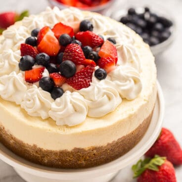 instant pot cheesecake on white cake plate with whipped cream strawberries and blueberries