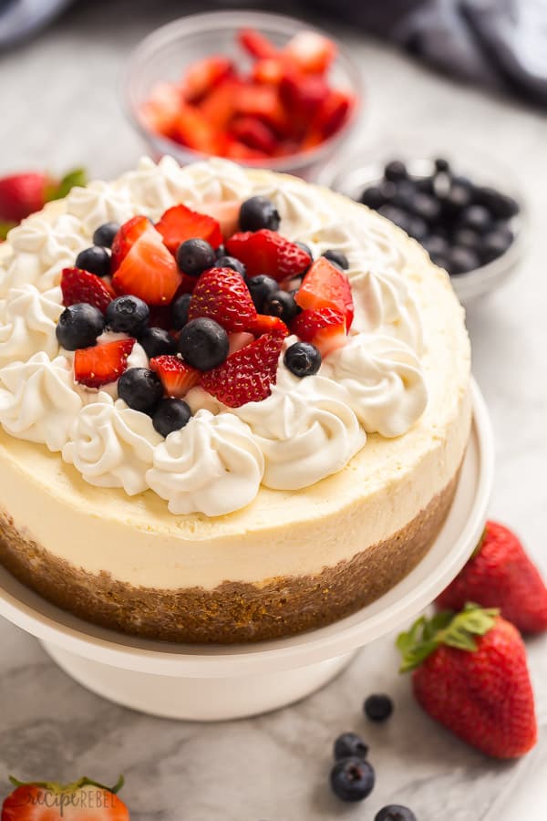 instant pot cheesecake on white cake plate with whipped cream strawberries and blueberries