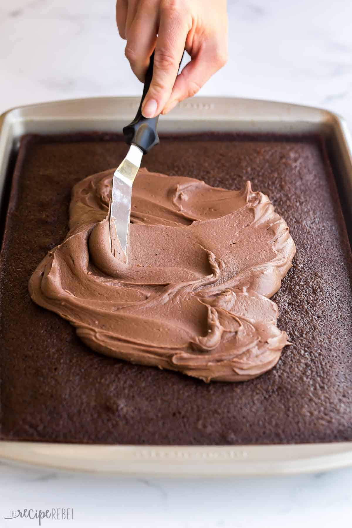 This Chocolate Sheet Cake is rich and chocolatey and so easy to make! Topped with a simple frosting made of melted butter, cocoa and sugar. It's sure to be a hit at any potluck! #chocolate #dessert #recipe | chocolate desserts | chocolate recipes | easy cake recipe | potluck dessert | holiday dessert | easy chocolate cake | easy dessert recipes spreading frosting on a chocolate sheet cake with an offset spatula.