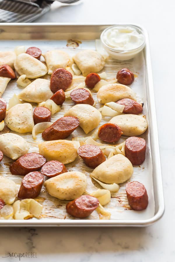 sliced sausages and perogies on a sheet pan with small bowl of sour cream