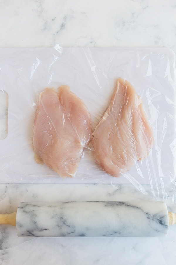 raw chicken breasts with plastic wrap over and rolling pin