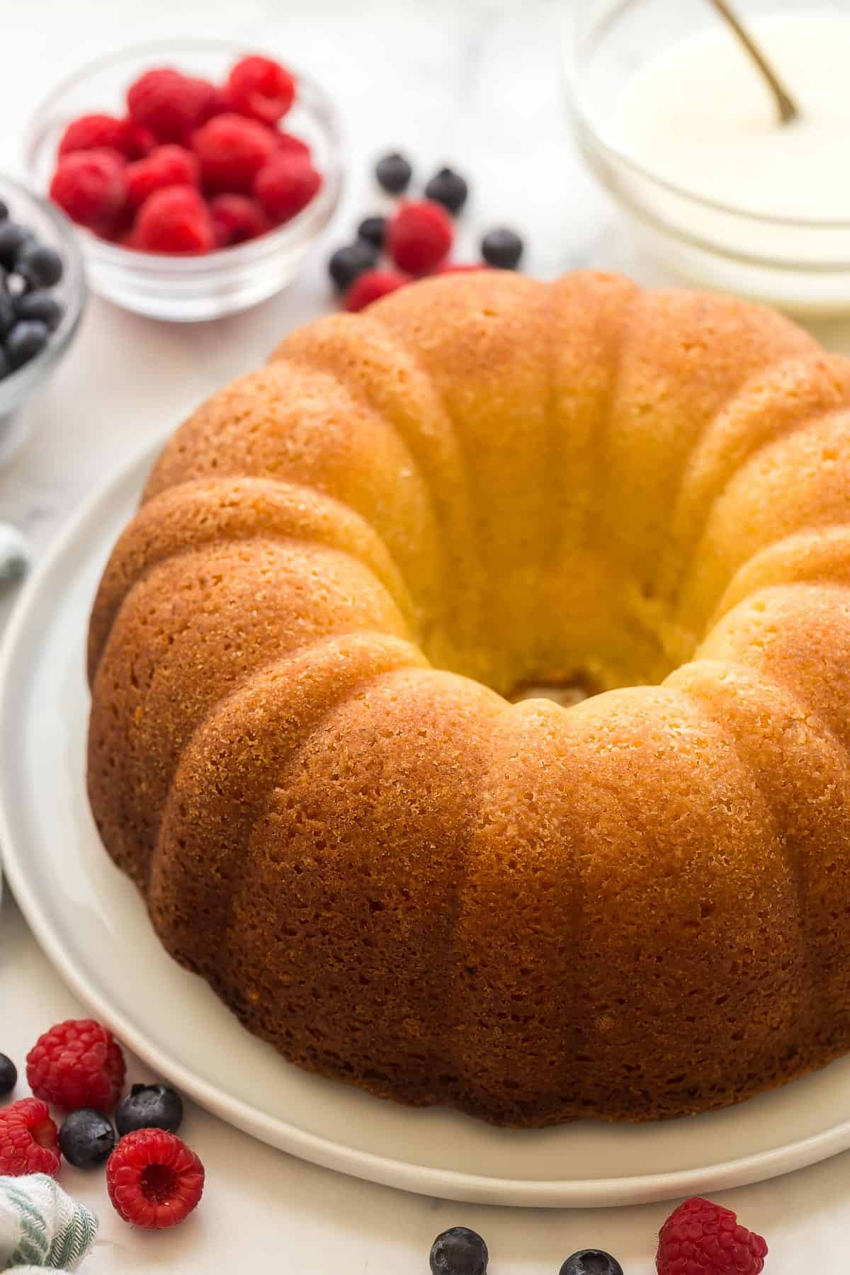 This Lemon Bundt Cake is so moist and packed with lemon flavor! It's full of real lemon flavor, topped with a lemon cream cheese glaze and berries. #lemon #cake #recipe | dessert recipes | lemon recipes | easy cake recipes | easter cake recipes | greek yogurt cake | easter treats | easter cake | spring desserts | bundt cakes whole lemon bundt cake without glaze on a white plate.