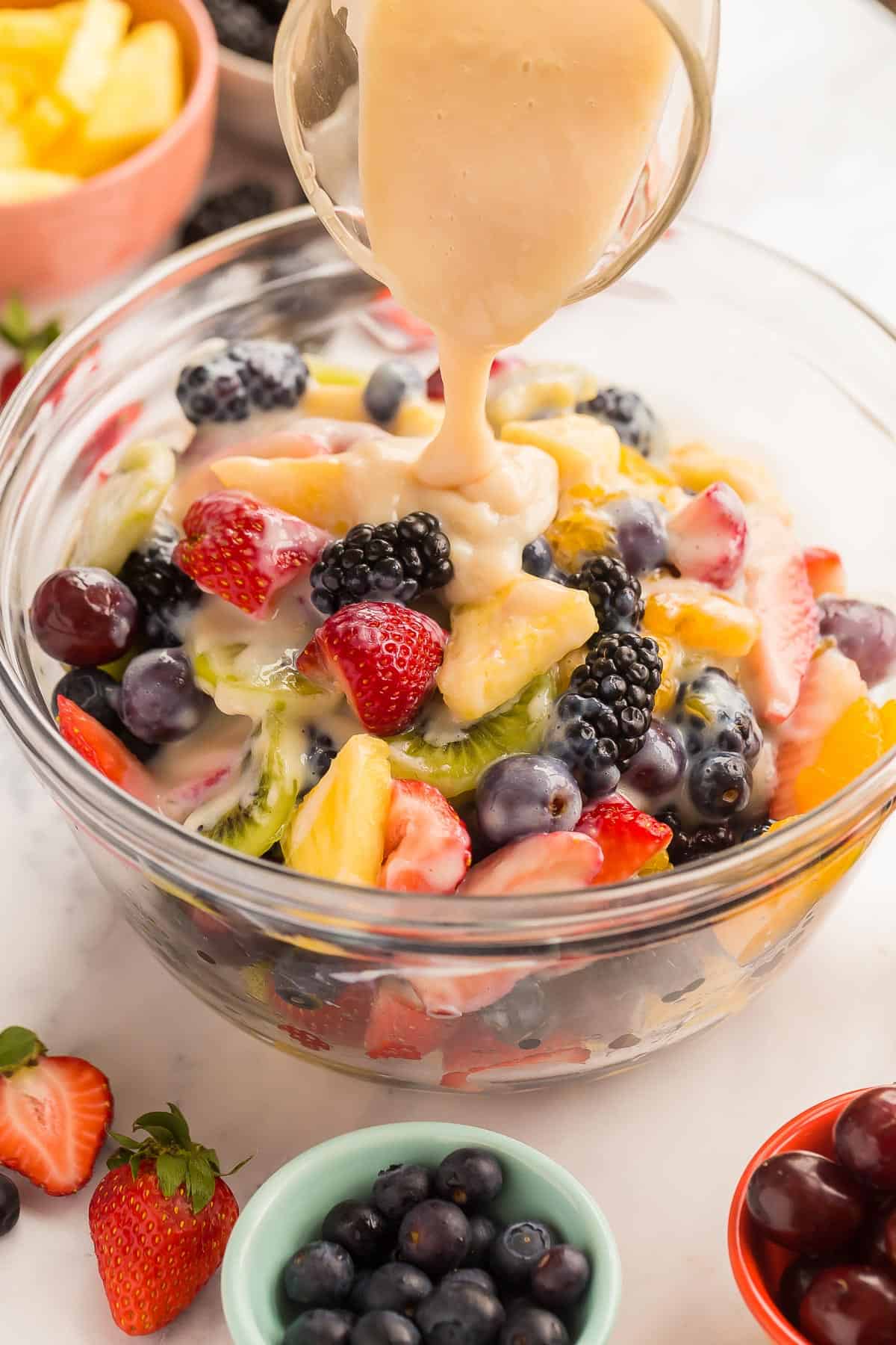 This Creamy Fruit Salad Recipe is made with a Homemade Vanilla Fruit Salad Dressing, plus strawberries, blueberries, oranges, grapes, kiwi and blackberries -- no pudding mix here! #fruit #salad #recipe | fruit salad recipe | homemade fruit salad dressing | berries | fresh fruit | spring dessert | easter brunch | easter dessert creamy vanilla dressing being poured on fruit salad in a glass bowl.