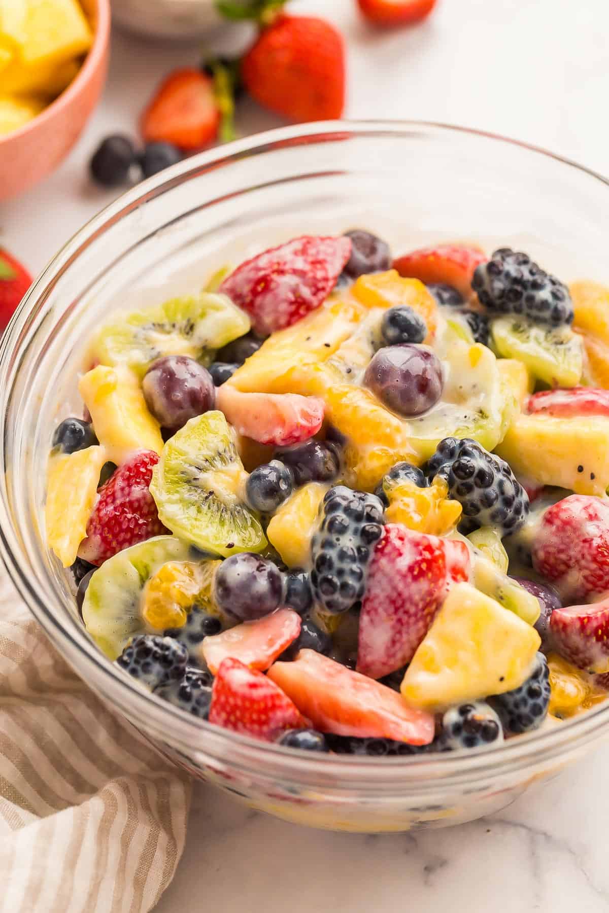 This Creamy Fruit Salad Recipe is made with a Homemade Vanilla Fruit Salad Dressing, plus strawberries, blueberries, oranges, grapes, kiwi and blackberries -- no pudding mix here! #fruit #salad #recipe | fruit salad recipe | homemade fruit salad dressing | berries | fresh fruit | spring dessert | easter brunch | easter dessert close up image of creamy fruit salad in a glass bowl with fresh fruit around.