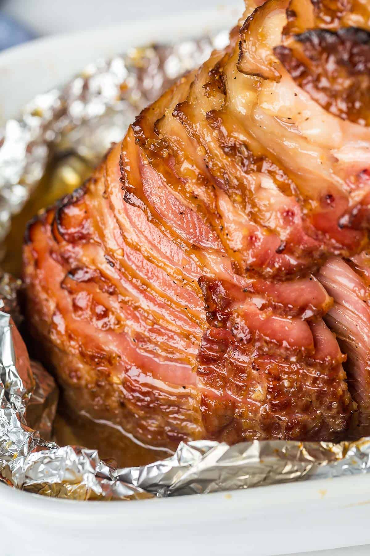 This Honey Baked Ham is coated in a sweet ham glaze, wrapped and baked until warmed through and full of flavor! The easiest way to get tender, juicy baked ham! #ham #easter #christmas #dinner | glazed ham | spiral ham | ham glaze | easter dinner | christmas dinner close up image of spiral ham with honey glaze in a baking dish.