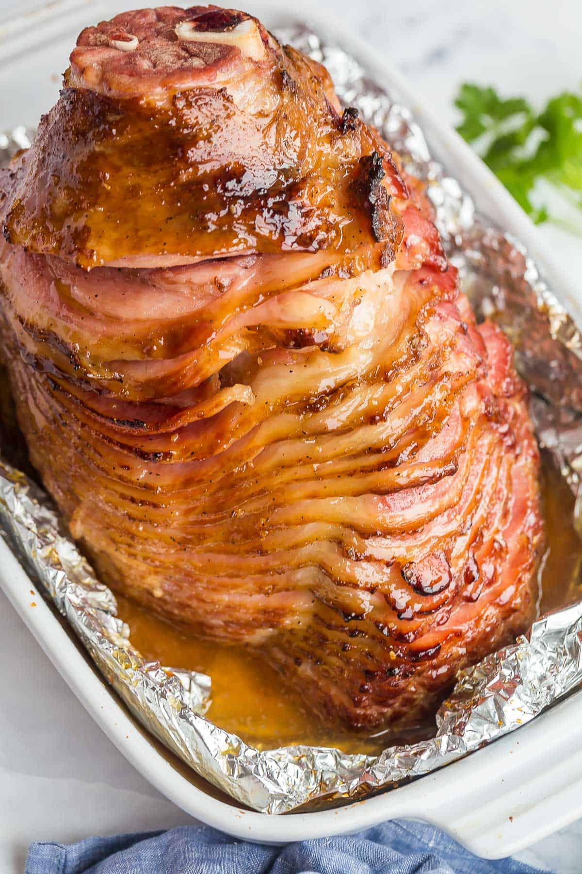 This Honey Baked Ham is coated in a sweet ham glaze, wrapped and baked until warmed through and full of flavor! The easiest way to get tender, juicy baked ham! #ham #easter #christmas #dinner | glazed ham | spiral ham | ham glaze | easter dinner | christmas dinner honey baked ham in white baking dish with foil around.