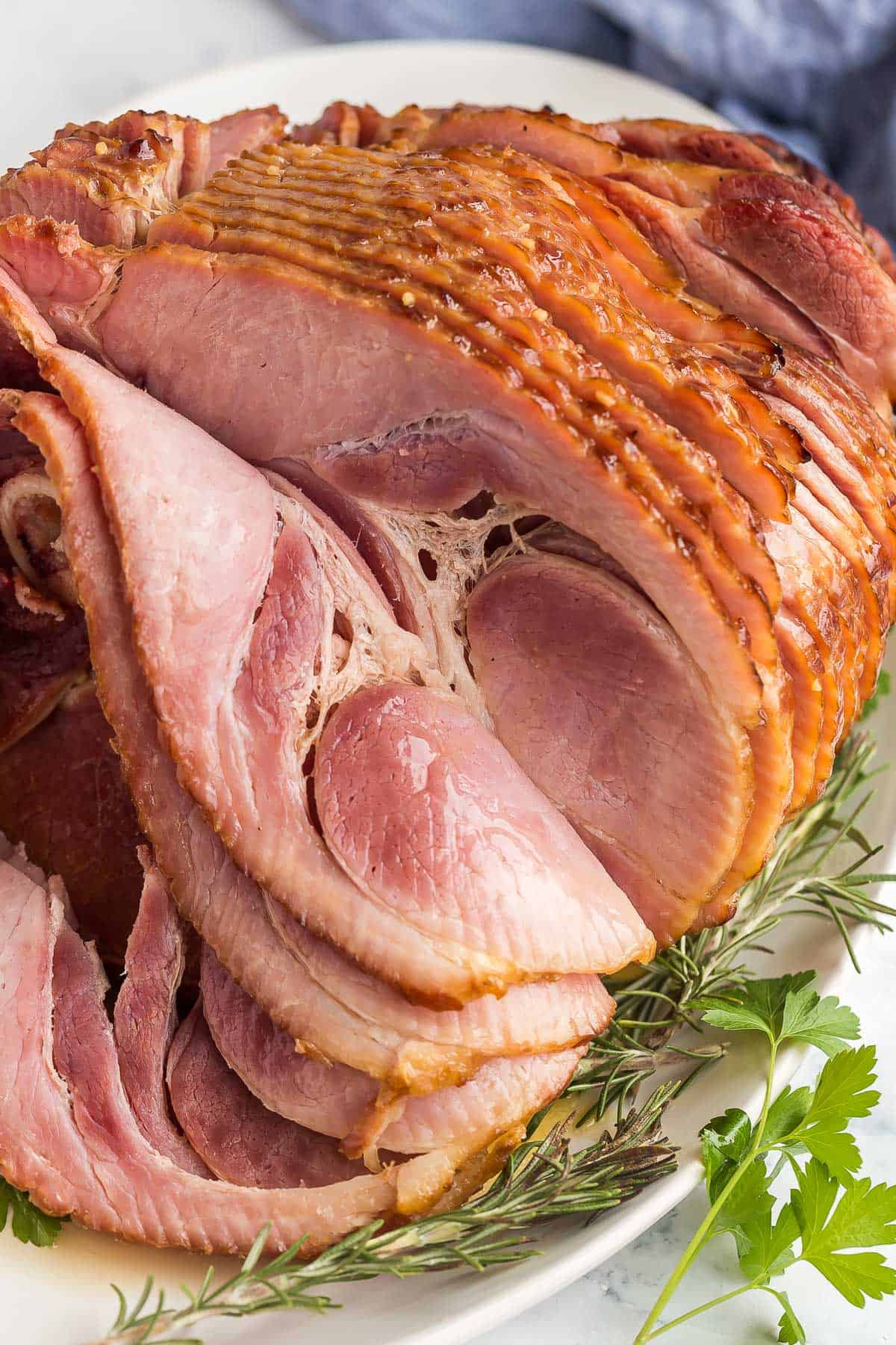 This Honey Baked Ham is coated in a sweet ham glaze, wrapped and baked until warmed through and full of flavor! The easiest way to get tender, juicy baked ham! #ham #easter #christmas #dinner | glazed ham | spiral ham | ham glaze | easter dinner | christmas dinner spiral ham on a white platter with parsley around.