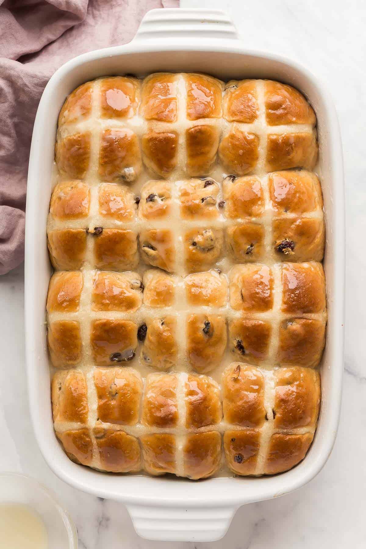 These Hot Cross Buns are sweet, fluffy, lightly spiced buns filled with raisins and marked with a white cross. They are traditionally served at Easter but delicious any time of year! #easter #baking #buns #recipes | easter recipes | easter baking | easter breakfast || easter brunch | cinnamon buns | cinnamon raisin buns | brunch recipes overhead image of hot cross buns in white baking dish.