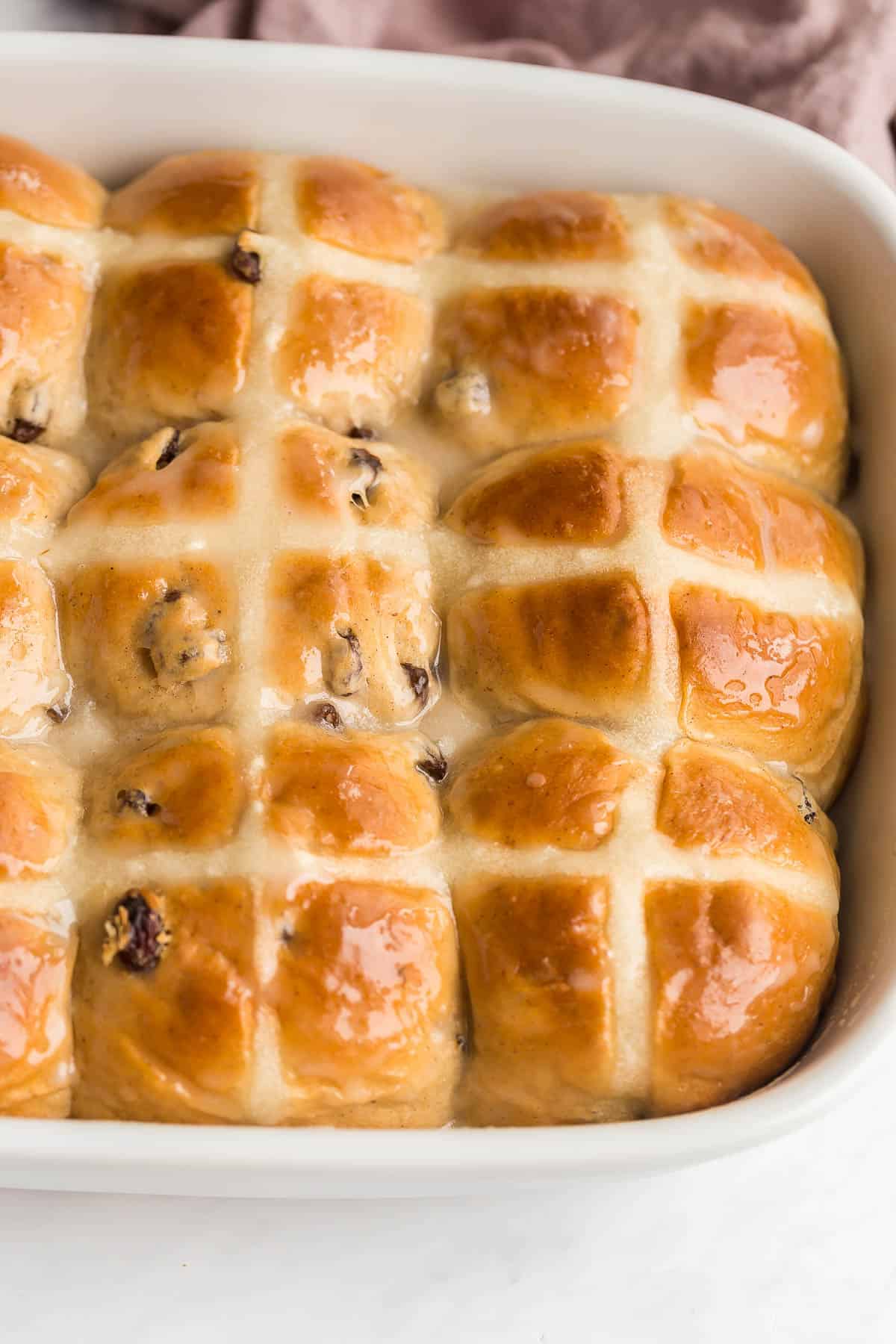These Hot Cross Buns are sweet, fluffy, lightly spiced buns filled with raisins and marked with a white cross. They are traditionally served at Easter but delicious any time of year! #easter #baking #buns #recipes | easter recipes | easter baking | easter breakfast || easter brunch | cinnamon buns | cinnamon raisin buns | brunch recipes close up image of some of the hot cross buns in a white baking dish.