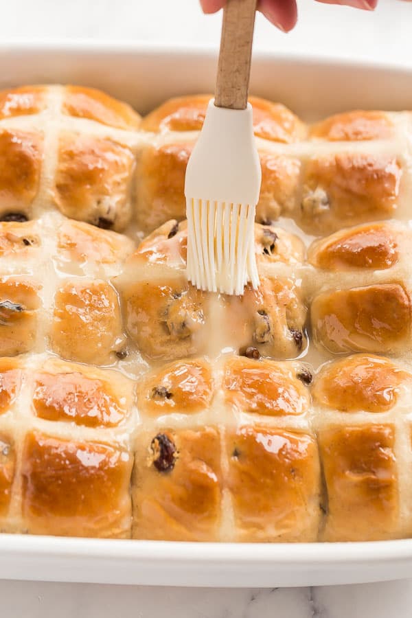 These Hot Cross Buns are sweet, fluffy, lightly spiced buns filled with raisins and marked with a white cross. They are traditionally served at Easter but delicious any time of year! #easter #baking #buns #recipes brushing glaze on hot cross buns