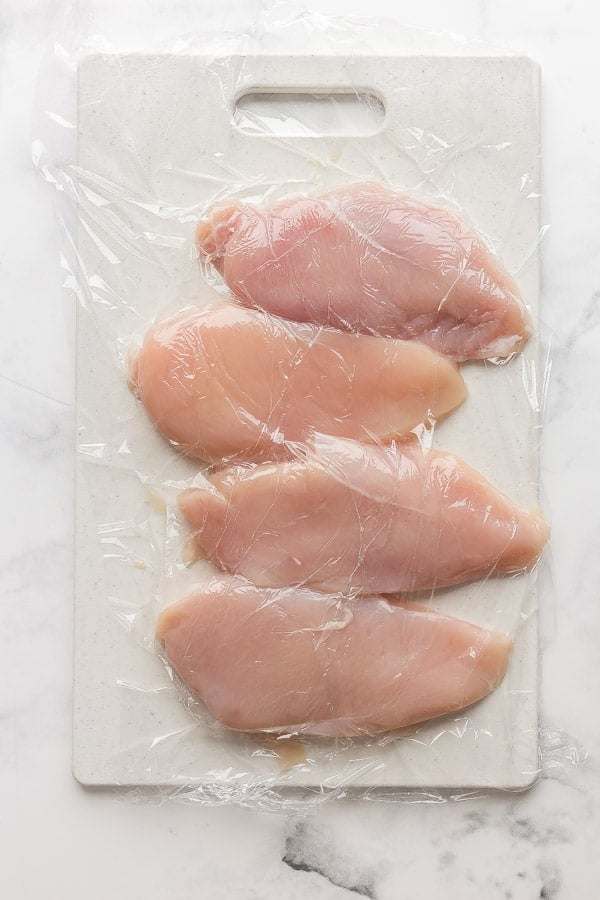 raw chicken breasts on cutting board with plastic wrap on top
