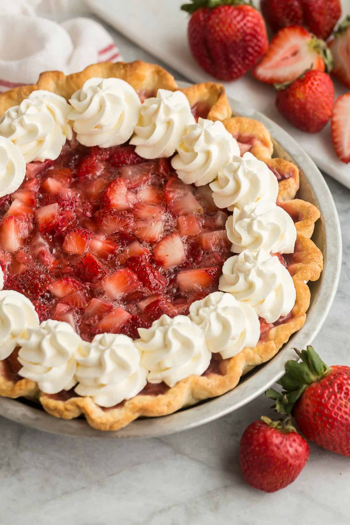 This fresh Strawberry Pie is made with real strawberries and no Jello mix in a homemade pie crust, topped with sweetened whipped cream! It's the perfect summer treat! #strawberry #dessert #recipe | dessert recipes | pie recipes | easy dessert ideas | summer dessert | strawberry recipes | summer recipes | easter pie close up image of fresh strawberry pie in tin pie plate with whipped cream.