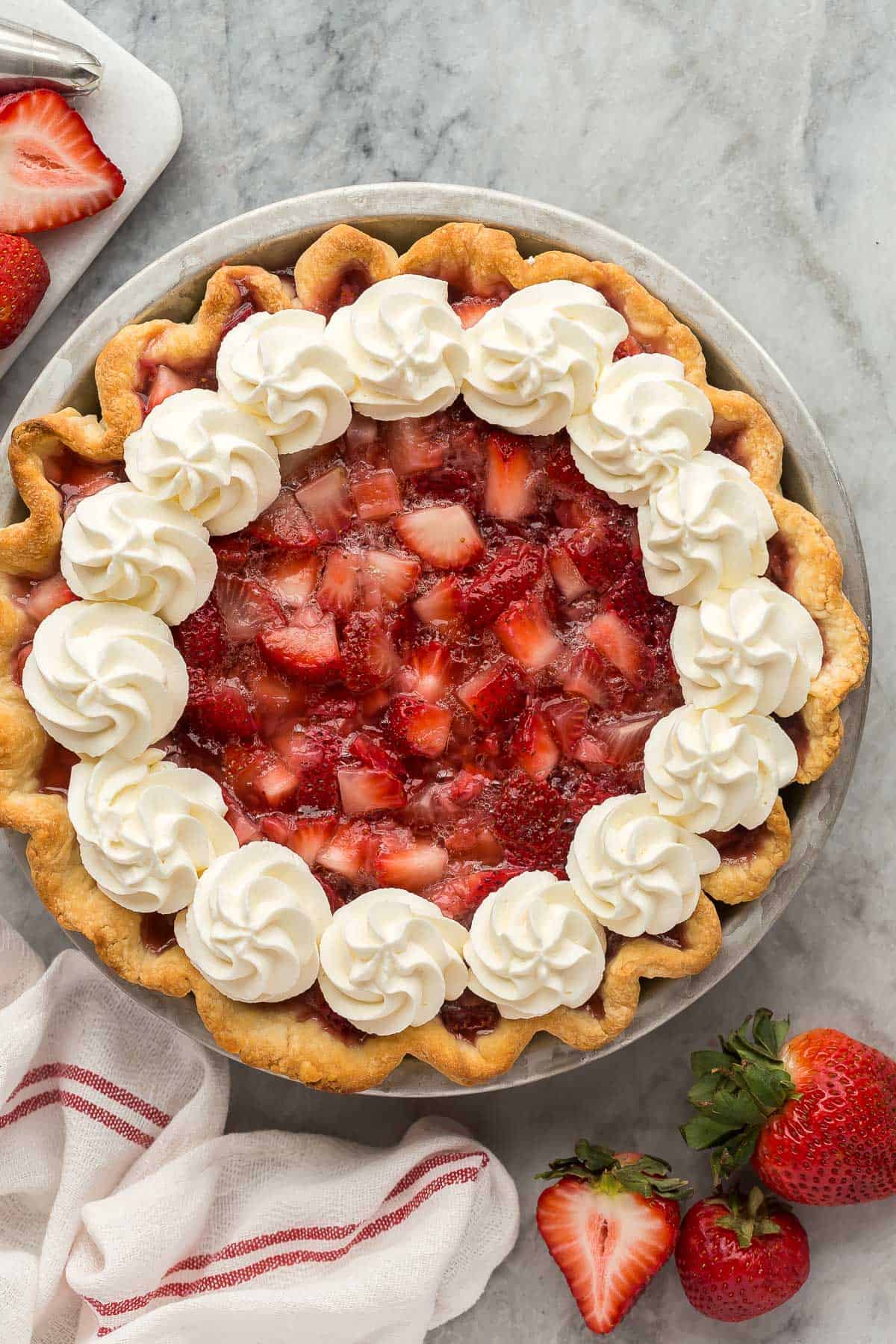 This fresh Strawberry Pie is made with real strawberries and no Jello mix in a homemade pie crust, topped with sweetened whipped cream! It's the perfect summer treat! #strawberry #dessert #recipe | dessert recipes | pie recipes | easy dessert ideas | summer dessert | strawberry recipes | summer recipes | easter pie overhead image of whole strawberry pie with whipped cream on marble background.