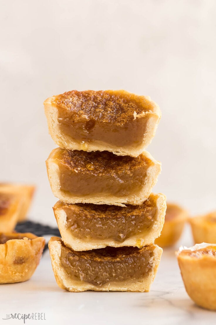 These Butter Tarts are a Canadian classic that comes together with just 25 minutes of prep! Homemade pastry shells are filled with a rich, brown sugar (raisins optional!), then baked until buttery and flaky on the inside and gooey on the inside. #baking #recipes | canadian recipes | tart shells | pie crust | homemade pastry | christmas treats | christmas baking | dessert recipes stack of four butter tarts cut in half to reveal filling