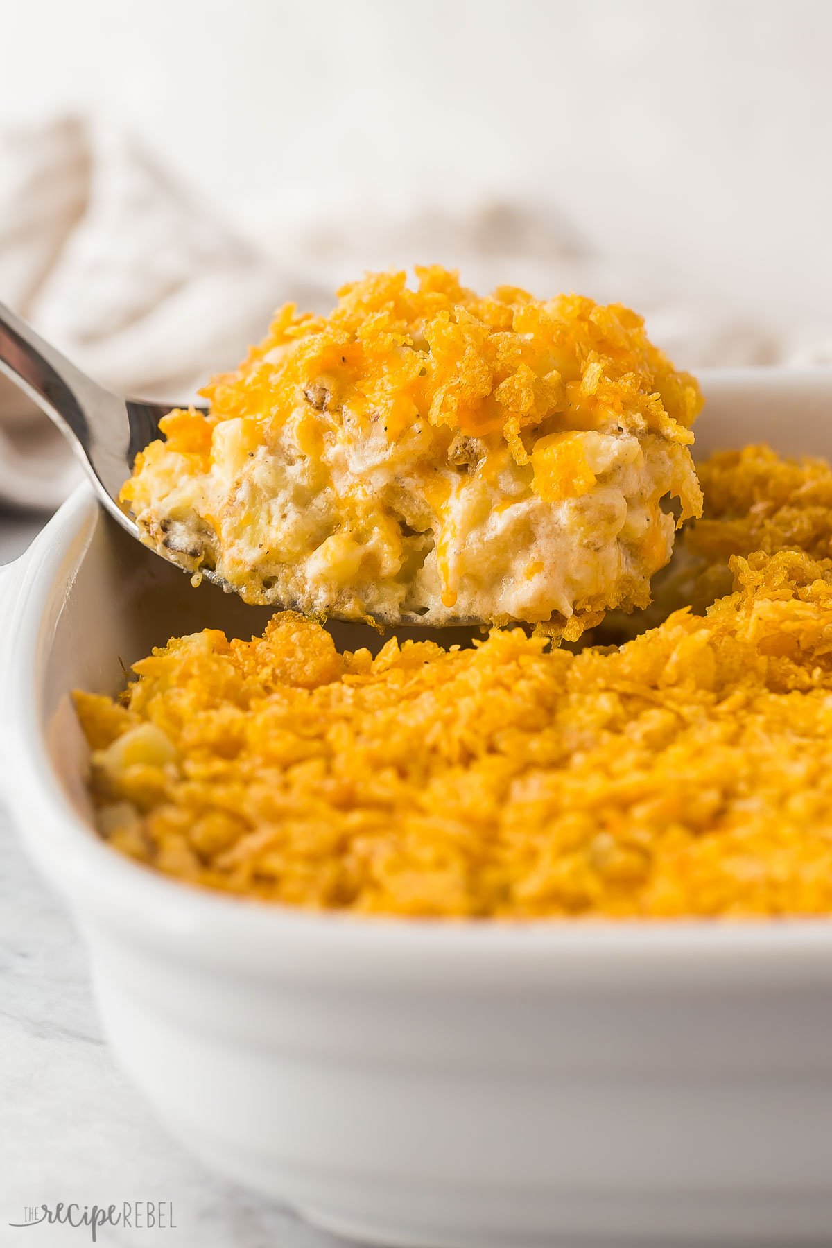 This Hashbrown Casserole is a classic side dish that takes 10 minute to prep! Hashbrown potatoes are baked in a rich and creamy sauce, topped with melty cheddar cheese, and finished off with a buttery, crunchy Corn Flake topping. #christsmas #thanksgiving #recipe | easy dinner ideas | thanksgiving dinner | christmas dinner | funeral potatoes | cheesy potatoes | easy recipes | frozen hashbrowns close up image of metal spoon scooping hashbrown casserole
