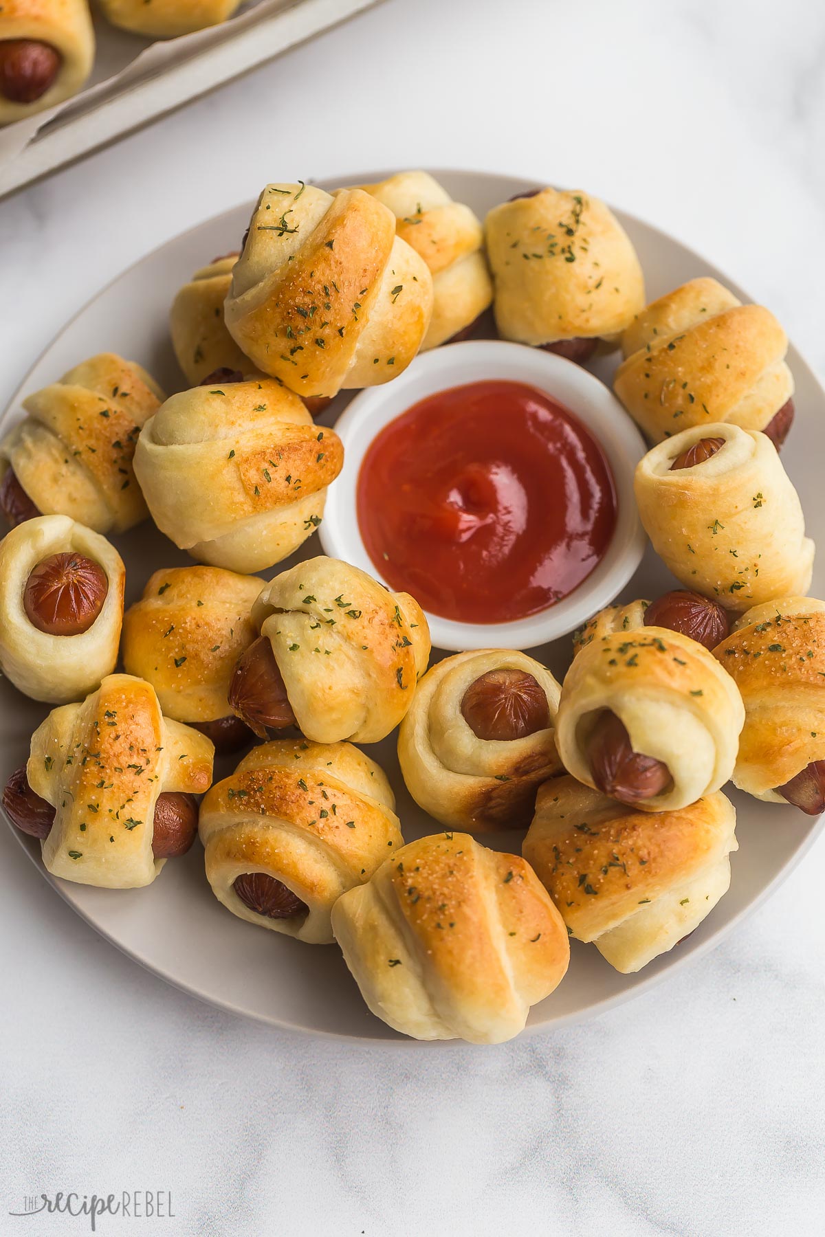 These Pigs in a Blanket are a completely homemade version of a classic. Little smokies are wrapped up in a simple homemade crescent dough, topped with flavorful seasonings, then baked until hot, golden-brown, and flaky. #appetizer #recipes #dinner | appetizer recipes | christmas recipe | dinner ideas | little smokies | kid friendly | holiday recipes | crescent dough recipes pigs in a blanket on a plate with bowl of ketchup in the middle