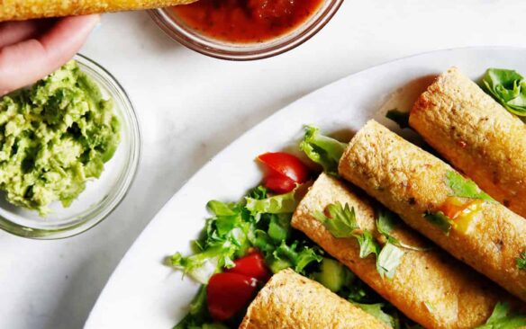 One chicken taquito is dipped into sauce next to a plate full of taquitos.