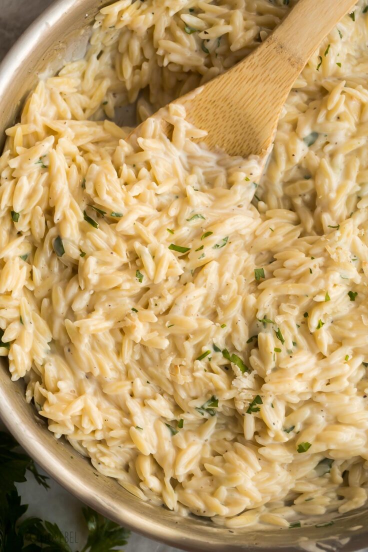 This Garlic Parmesan Orzo Pasta is a one-pot recipe for tender orzo tossed in a light and cheesy cream sauce. It's simple to make and comes together in a total of 20 minutes! #pasta #recipes #dinner | easy dinner ideas | dinner recipes | easy recipe | pasta recipe | orzo pasta | garlic | cheesy | comfort food close up image of creamy orzo pasta in skillet with wooden spoon
