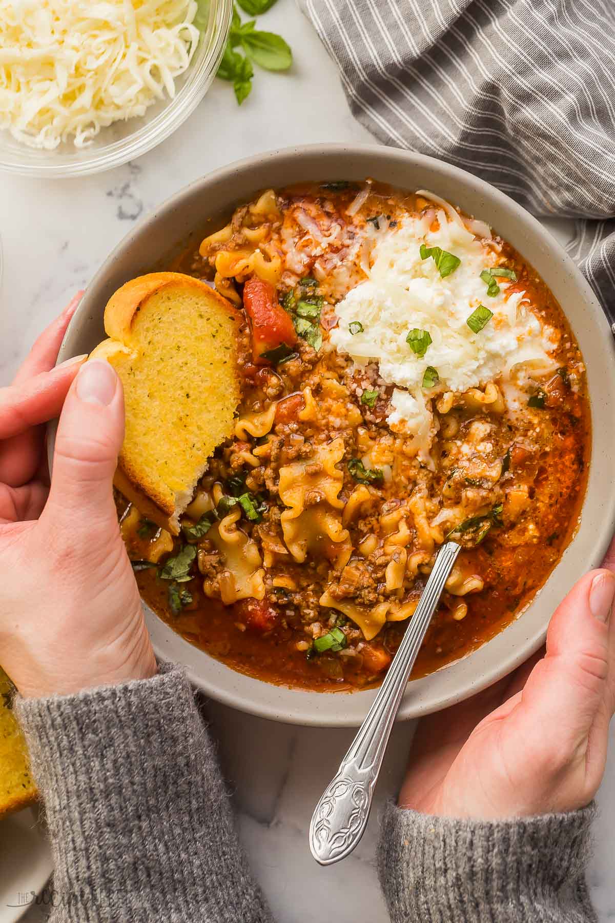 overhead image of two hands holding bowl of lasagna soup with piece of garlic bread in one hand